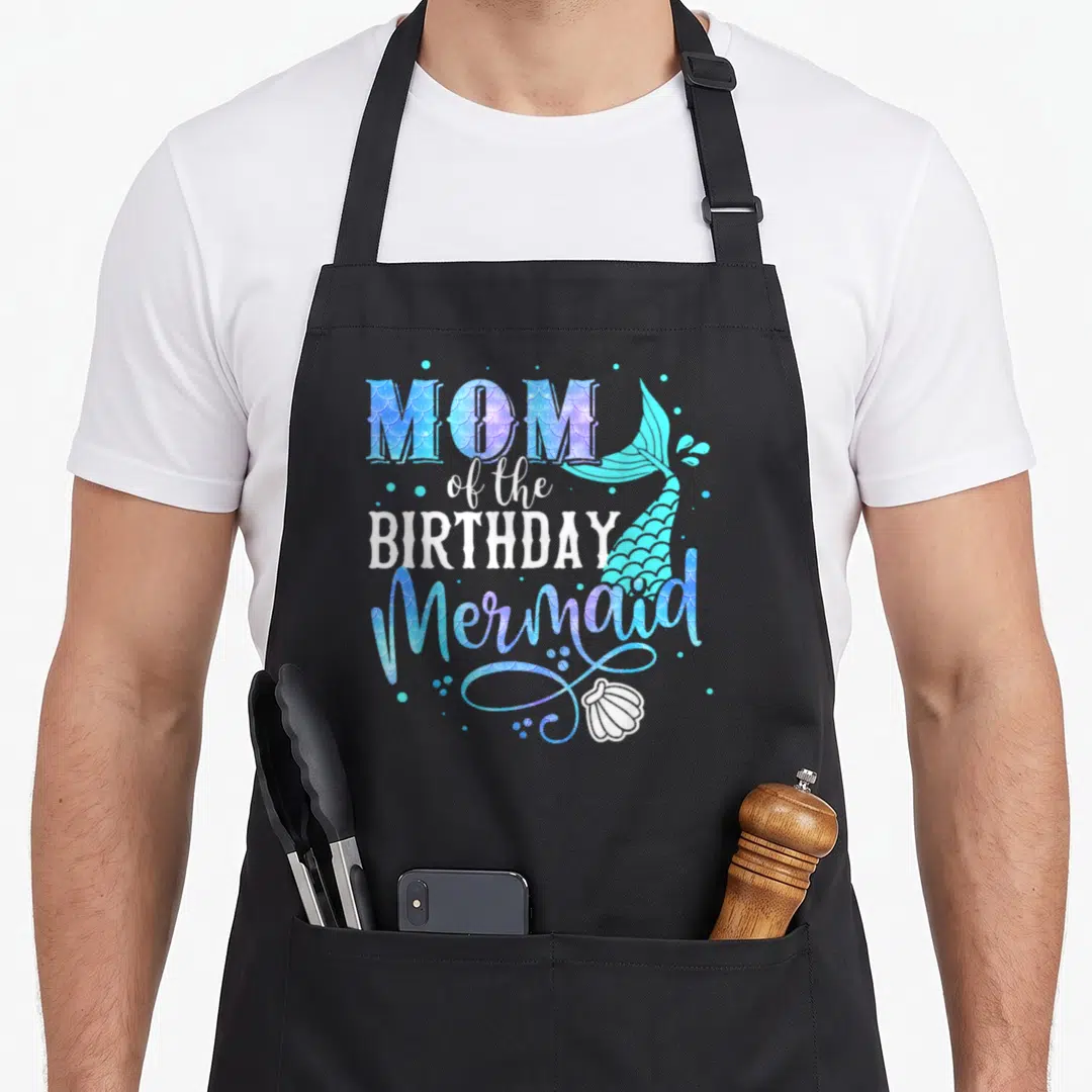 Family mother's apron - MOM of the BIRTHDAY Mermaid text with blue tail and seashell — shown on a black cotton canvas apron against a white background