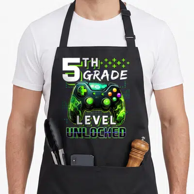 School level apron - 5th Grade Level Unlocked game controller print with green plus signs and blue grid background — shown on a black cotton canvas apron against a white background