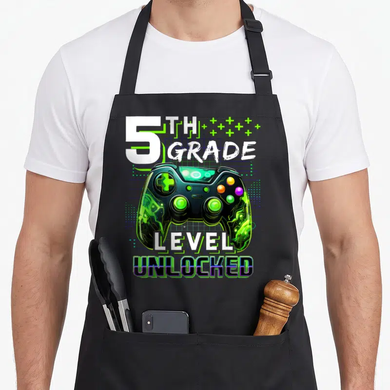 School level apron - 5th Grade Level Unlocked game controller print with green plus signs and blue grid background — shown on a black cotton canvas apron against a white background