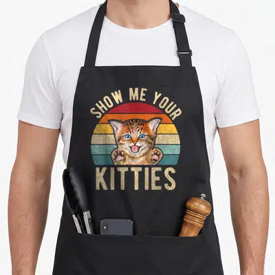 Show kitten kitty cat apron - Retro striped sun with blue-eyed kitten and SHOW ME YOUR KITTIES text in beige — shown on a black cotton canvas apron against a white background