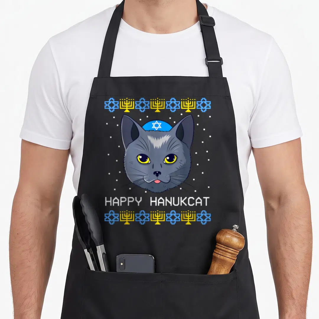 Hanukcat jewish apron - Grey cartoon cat head wearing blue kippah with white text HAPPY HANUKCAT flanked by yellow — shown on a black cotton canvas apron against a white background