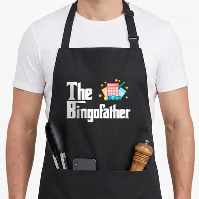 Lucky bingo aprons - White text The Bingofather next to red blue yellow bingo cards and green marker — shown on a black cotton canvas apron against a white background