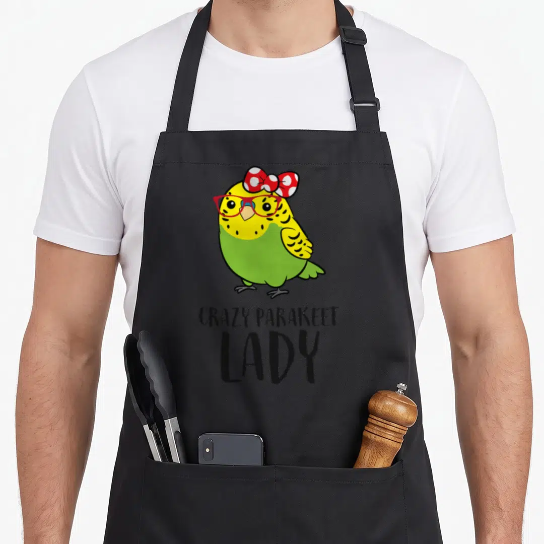 Comfort bird apron - Cartoon parakeet with red glasses and bow above stacked text CRAZY PARAKEET LADY — shown on a black cotton canvas apron against a white background
