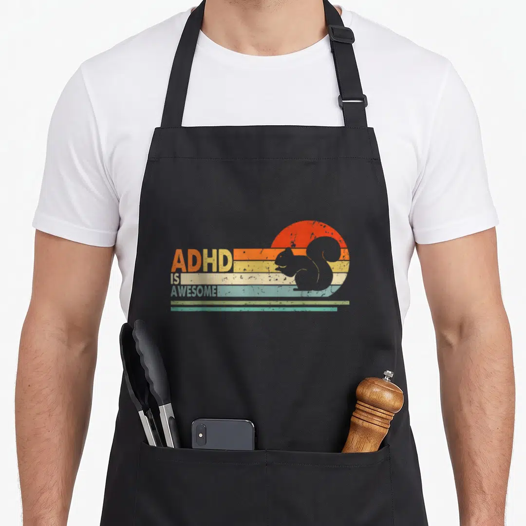 Vintage adhd squirrel aprons - ADHD IS AWESOME! text with squirrel silhouette on retro striped sun in orange red yellow — shown on a black cotton canvas apron against a white background