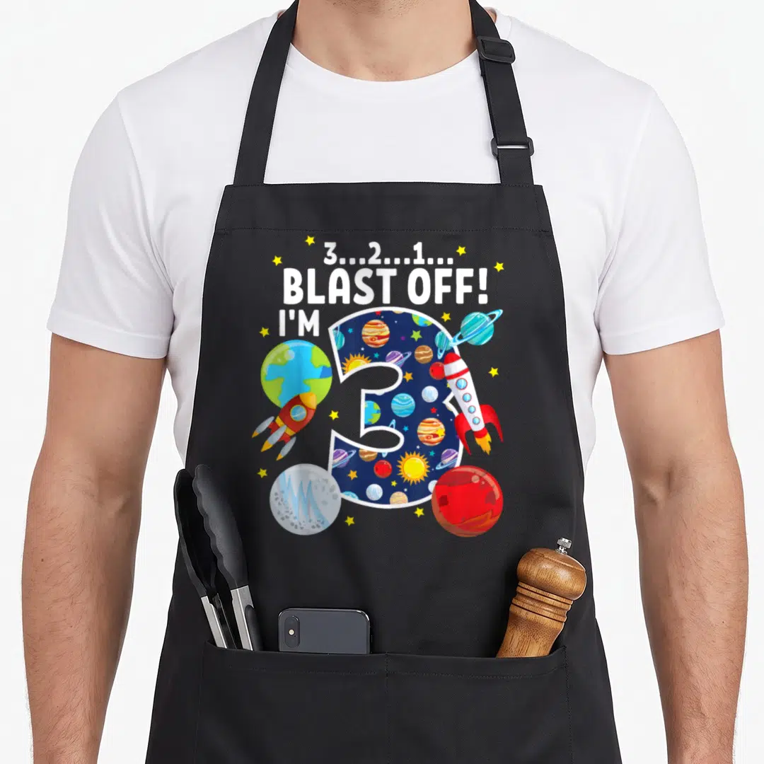 Space blast birthday apron - Number 3 filled with space pattern surrounded by rockets planets and blast off text — shown on a black cotton canvas apron against a white background