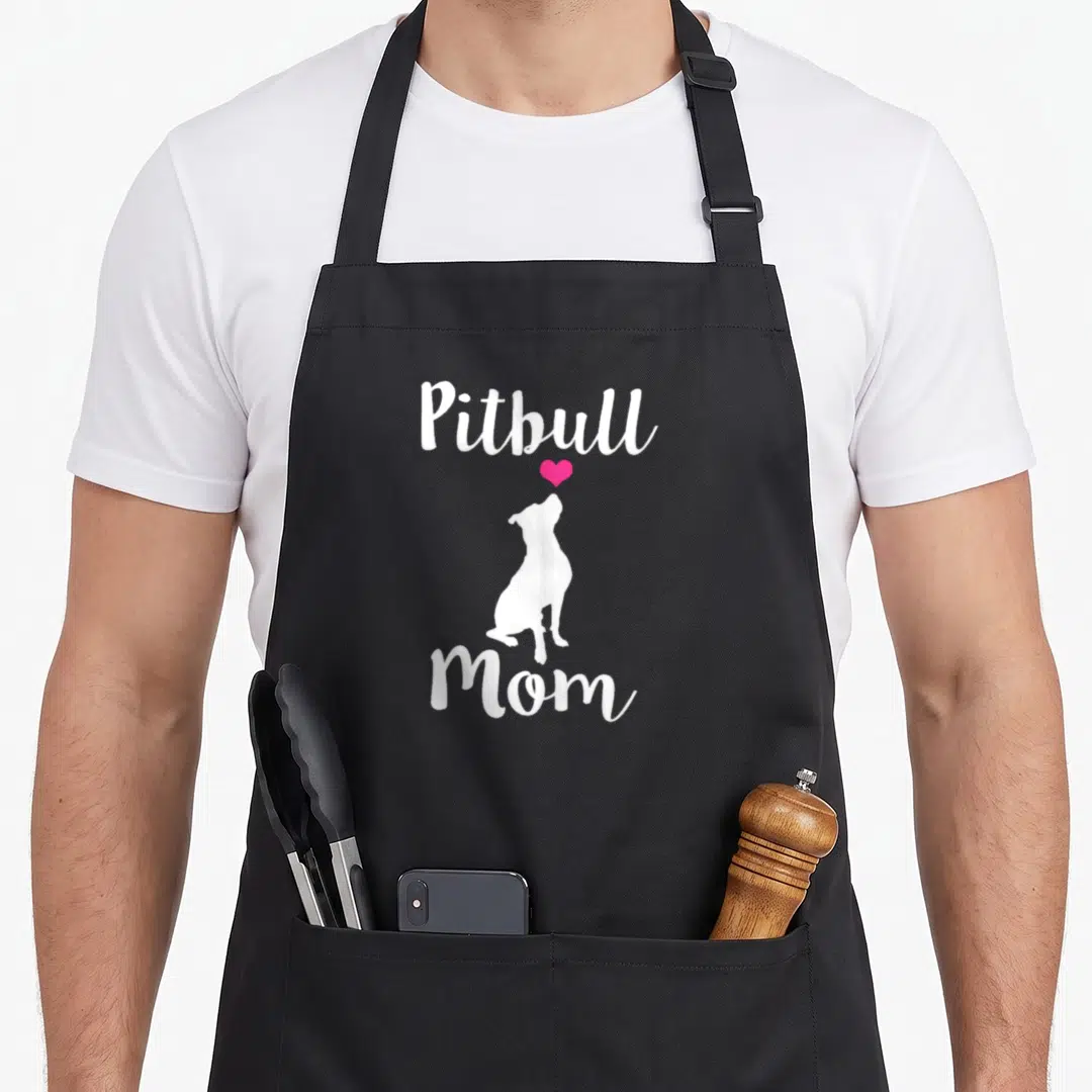 Pitbull pit dog apron - White Pitbull Mom text with sitting dog silhouette and pink heart — shown on a black cotton canvas apron against a white background
