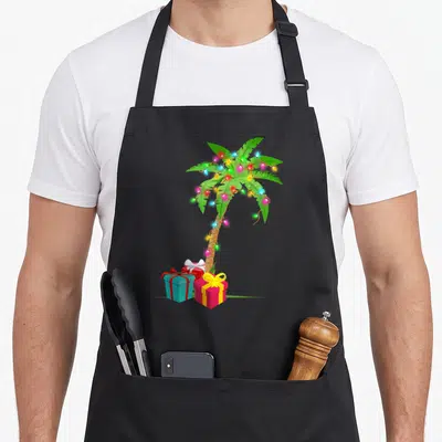 Coconut christmas tree apron - Cartoon palm tree with multicolor lights and three gift boxes on grass — shown on a black cotton canvas apron against a white background