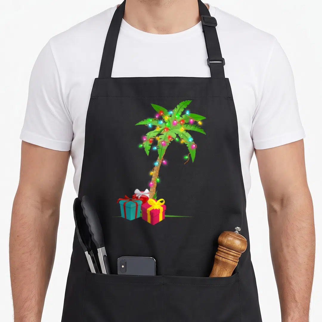 Coconut christmas tree apron - Cartoon palm tree with multicolor lights and three gift boxes on grass — shown on a black cotton canvas apron against a white background