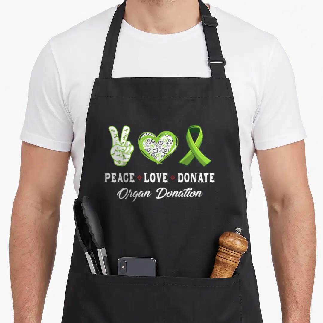 Peace organ apron - Peace sign hand, patterned heart, green ribbon, and stacked text reading PEACE LOVE DONATE Organ — shown on a black cotton canvas apron against a white background