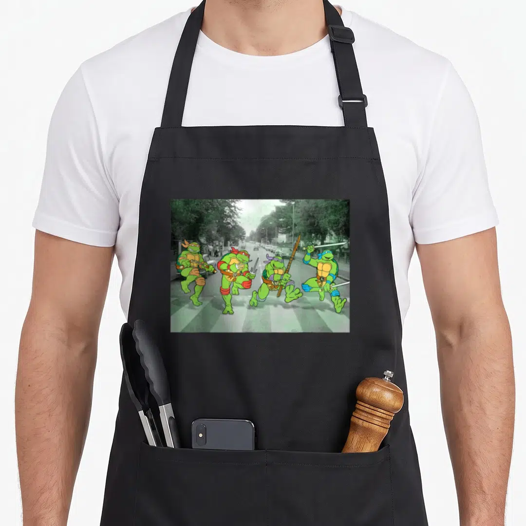 Parody road apron - Four green turtles with weapons walking across a striped crosswalk past grey cars and trees — shown on a black cotton canvas apron against a white background