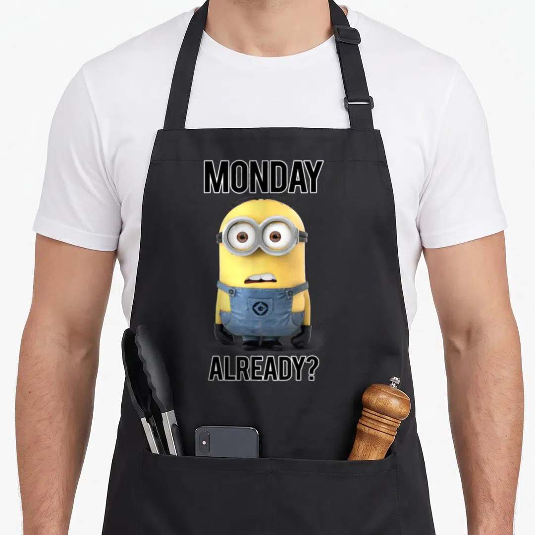 Despicable crew neck apron - Shocked yellow Minion wearing blue overalls with Monday Already text above and below — shown on a black cotton canvas apron against a white background
