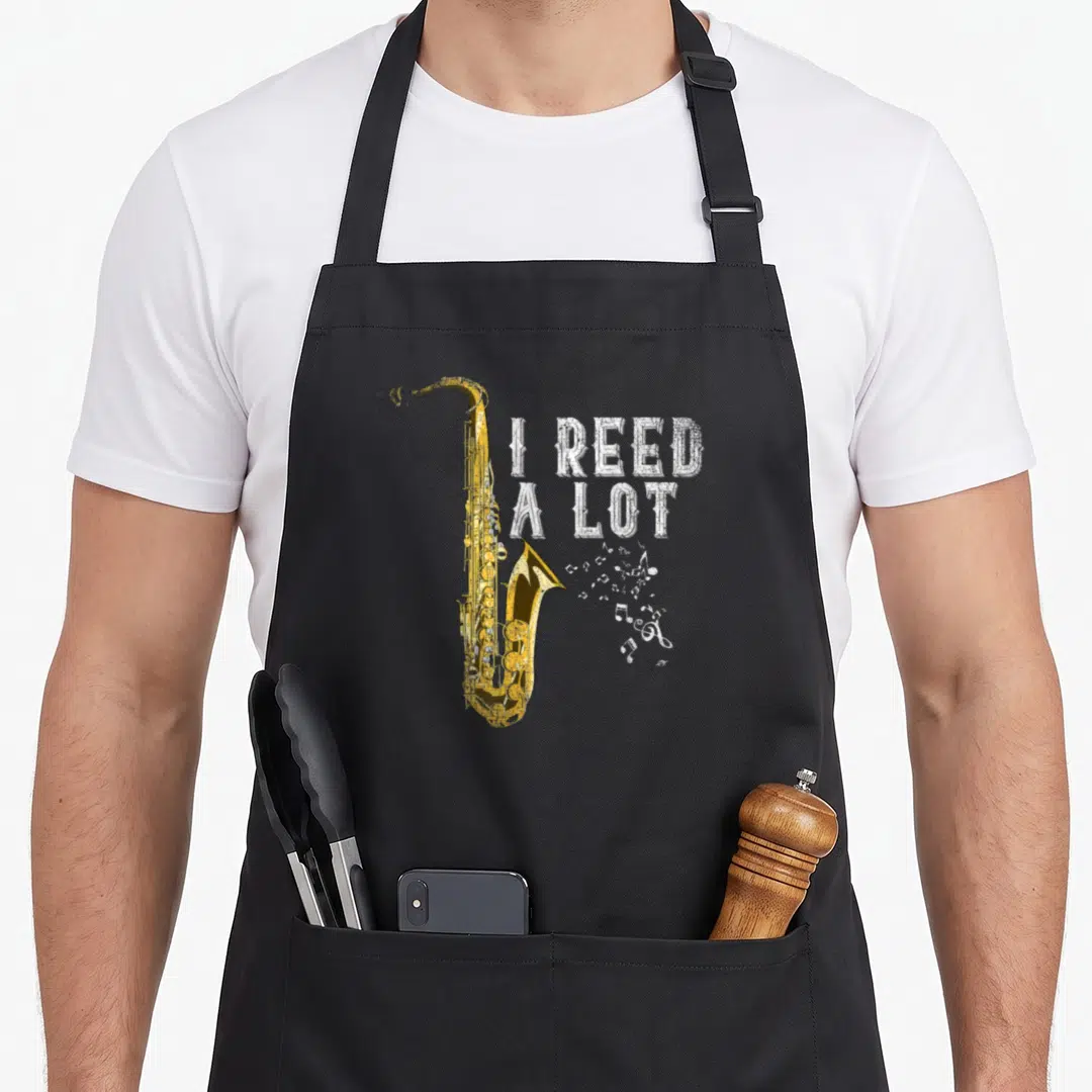 Musician saxophonist apron - Gold saxophone illustration beside white distressed text I REED A LOT with scattered — shown on a black cotton canvas apron against a white background