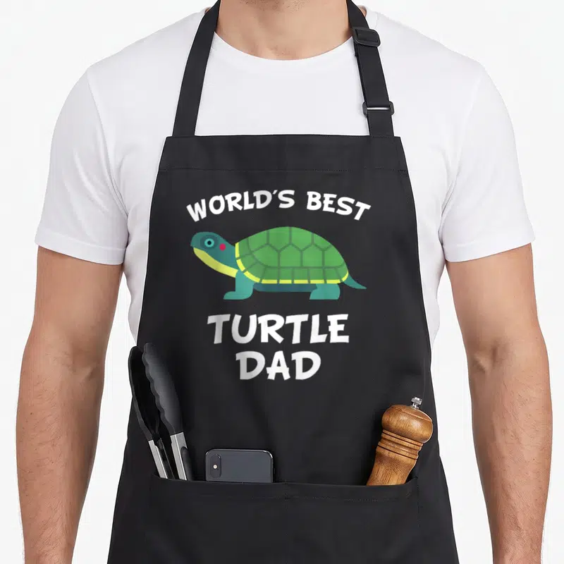 Owner turtle dad apron - Cartoon green turtle with white WORLD'S BEST TURTLE DAD text — shown on a black cotton canvas apron against a white background