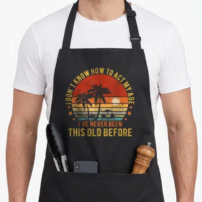 Funny general act age apron - Retro sunset with palm trees and text I don't know how to act my age — shown on a black cotton canvas apron against a white background