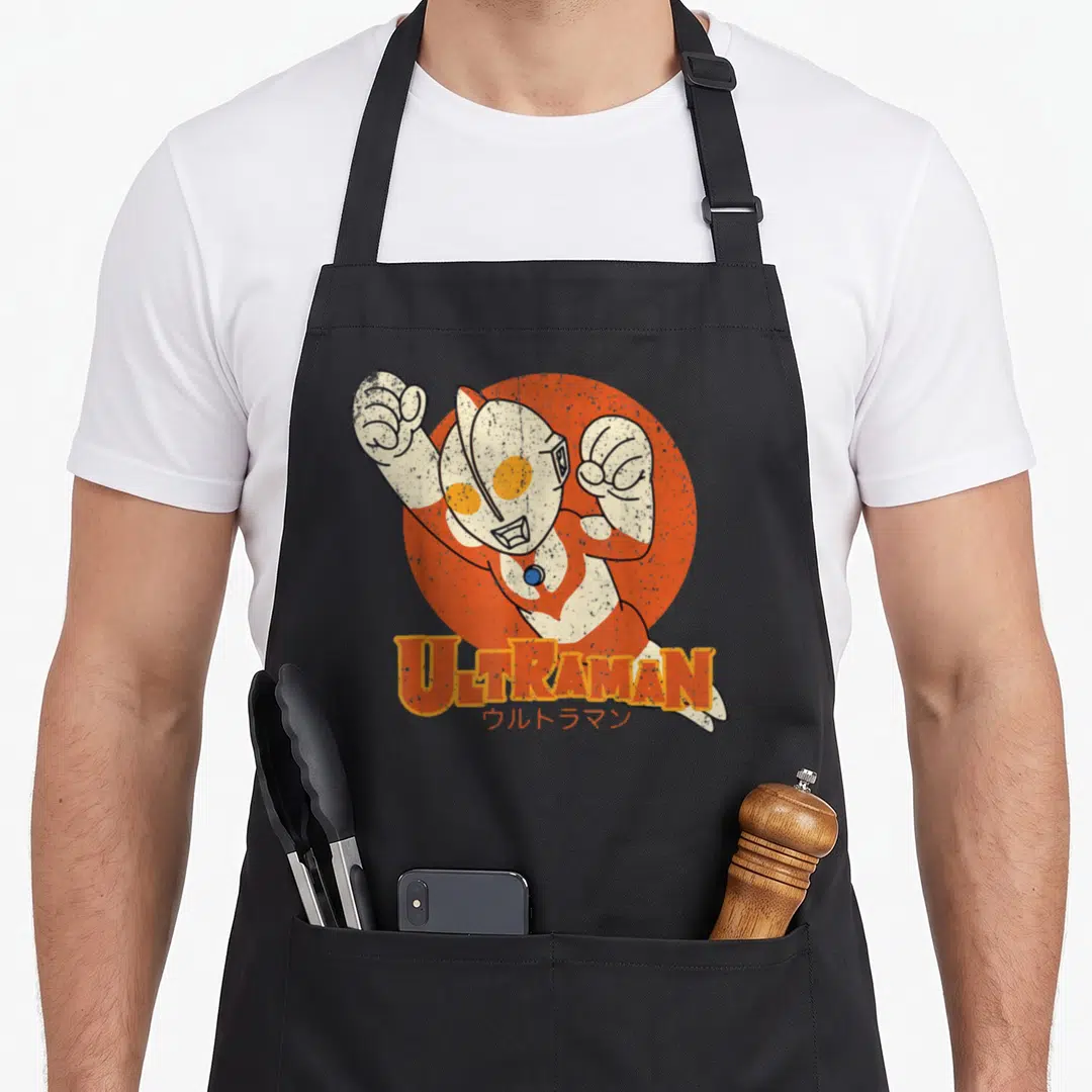 Ultraman kawaii apron - Chibi Ultraman cartoon over orange circle with stacked English and Japanese text — shown on a black cotton canvas apron against a white background