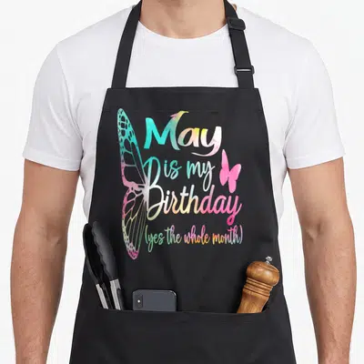Cool may birthday apron - Stacked watercolor text May is my Birthday with large left wing and small right wing in pink — shown on a black cotton canvas apron against a white background