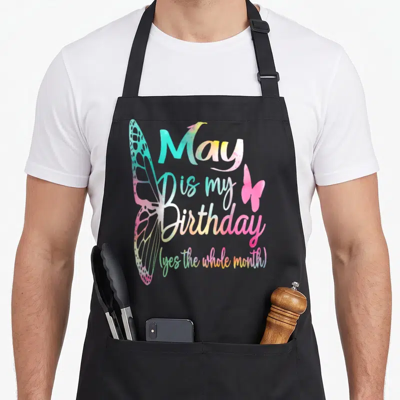 Cool may birthday apron - Stacked watercolor text May is my Birthday with large left wing and small right wing in pink — shown on a black cotton canvas apron against a white background
