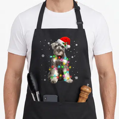 Sweater schnauzer apron - Grey dog wearing red santa hat wrapped in colorful christmas lights with white snowflakes — shown on a black cotton canvas apron against a white background