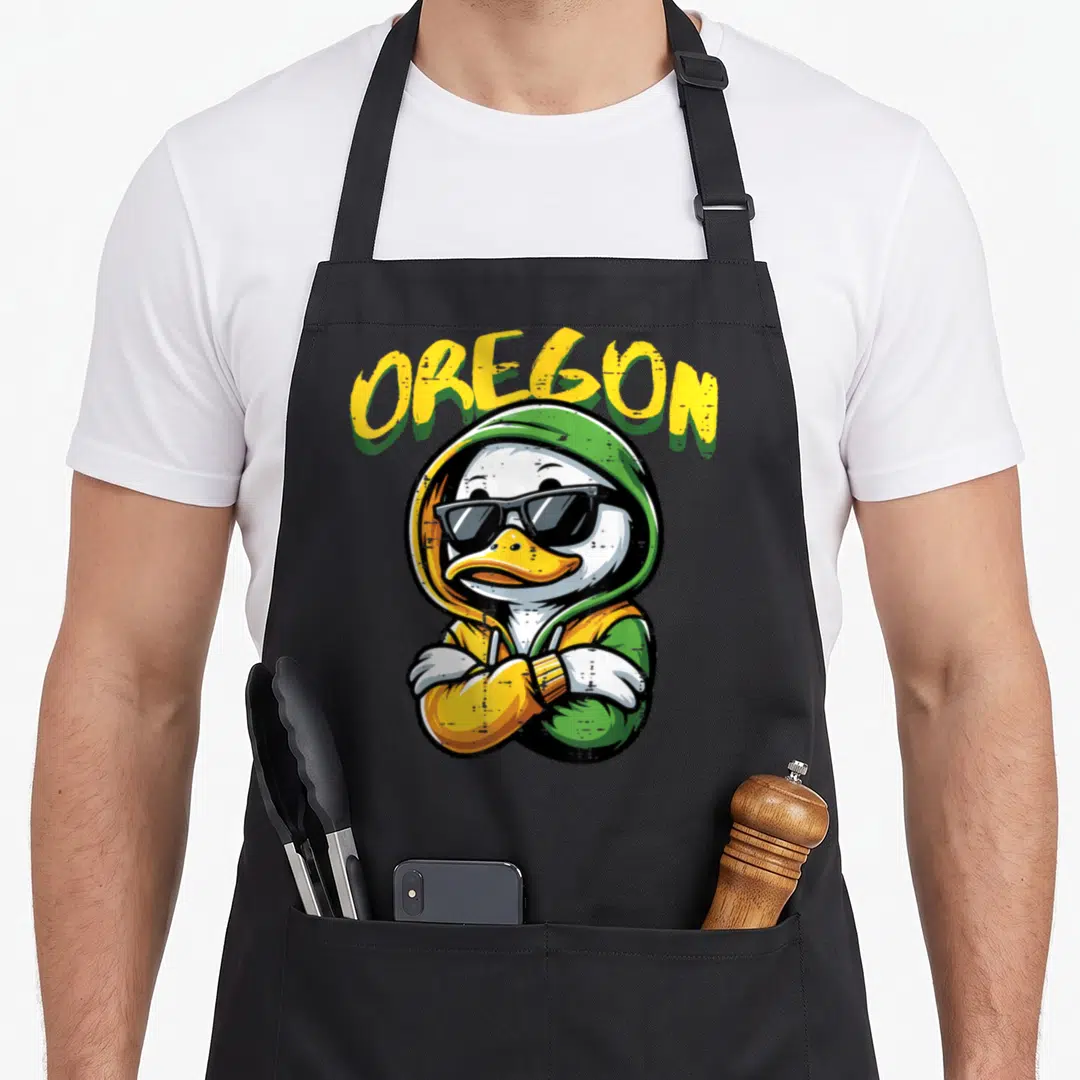 Sunglass duck apron - cartoon duck in black sunglasses and green yellow hoodie under arched OREGON text — shown on a black cotton canvas apron against a white background