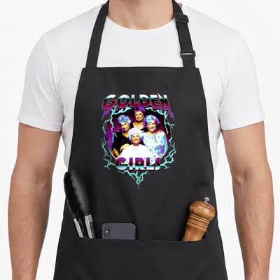 80s golden apron - GOLDEN GIRLS text with four women and electric bolts in purple teal blue retro style — shown on a black cotton canvas apron against a white background