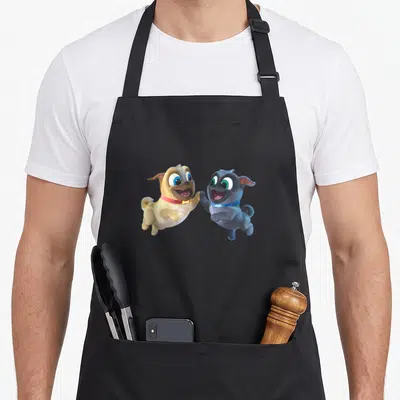 Pal dog apron - Two cartoon puppies jumping to high-five in tan, grey, and blue on black fabric — shown on a black cotton canvas apron against a white background
