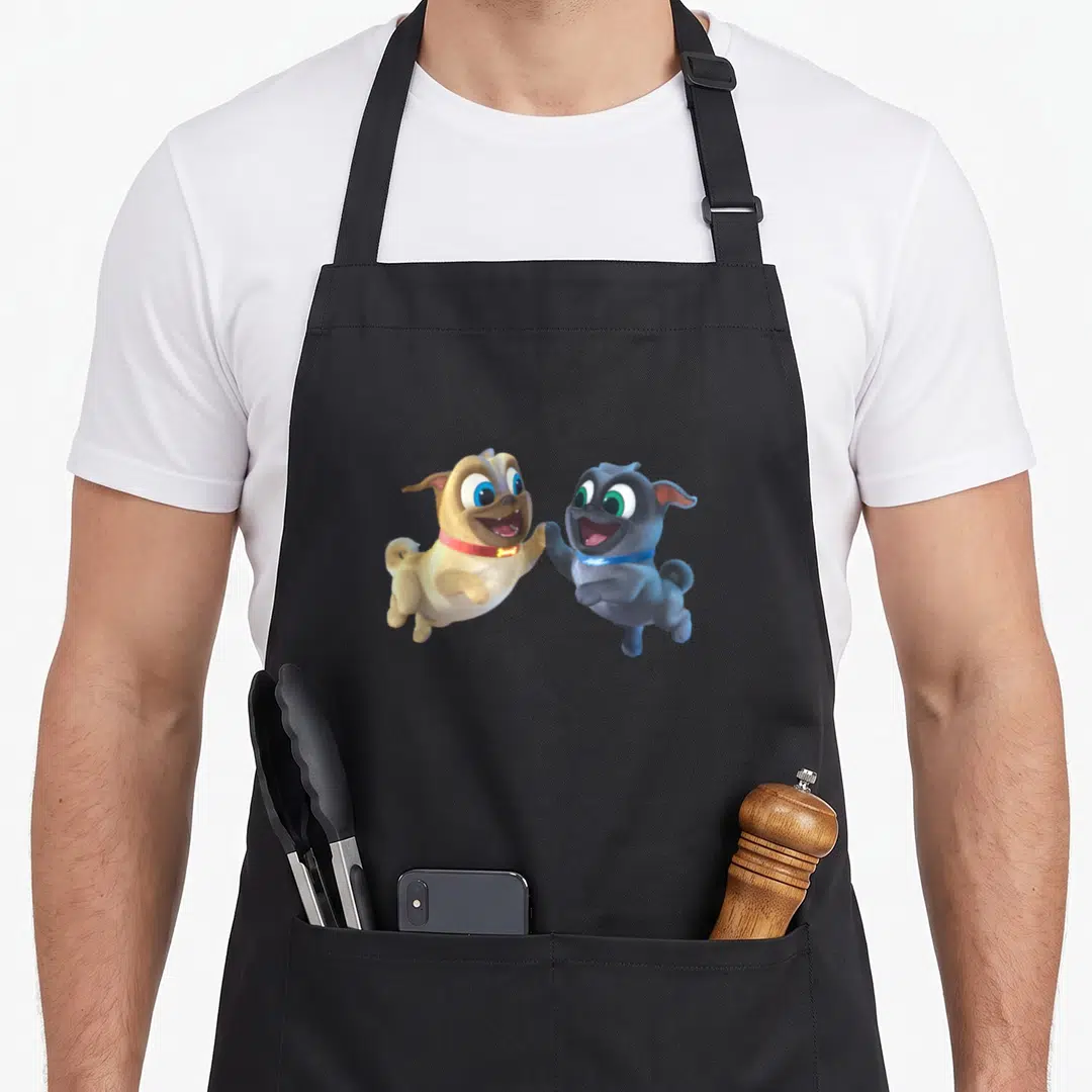 Pal dog apron - Two cartoon puppies jumping to high-five in tan, grey, and blue on black fabric — shown on a black cotton canvas apron against a white background