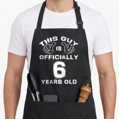 Officially 6th birthday apron - White pointing hands and text reading This Guy Is Officially 6 Years Old — shown on a black cotton canvas apron against a white background