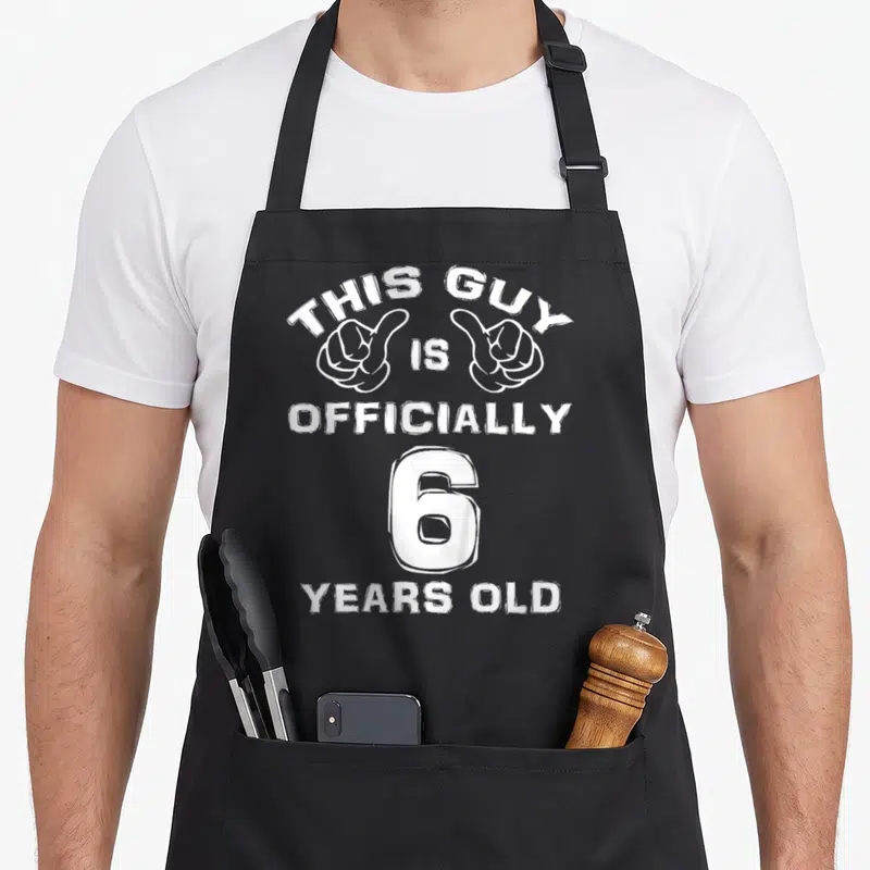 Officially 6th birthday apron - White pointing hands and text reading This Guy Is Officially 6 Years Old — shown on a black cotton canvas apron against a white background