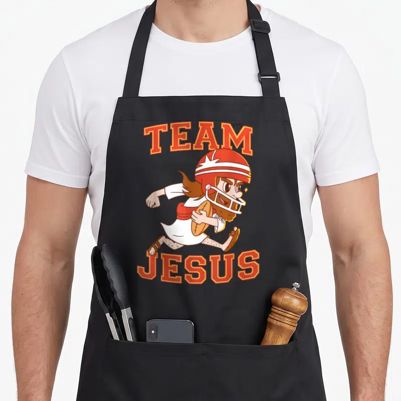 Christian football team aprons - Cartoon Jesus running in helmet holding football with TEAM JESUS text in red — shown on a black cotton canvas apron against a white background