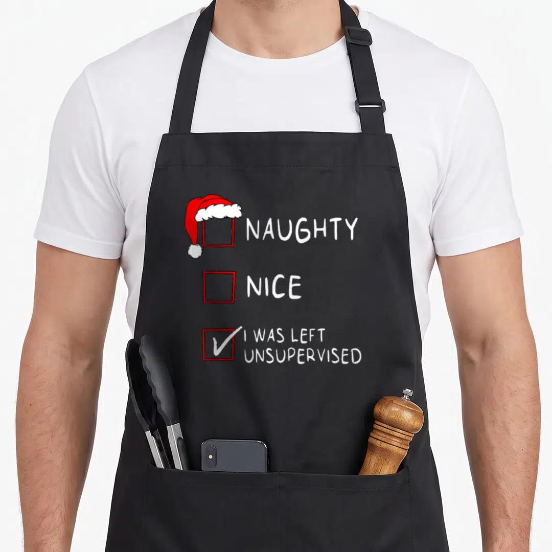 Naughty left apron - Cartoon checklist with red boxes, white text, Santa hat, and checkmark reading Naughty Nice I Was — shown on a black cotton canvas apron against a white background