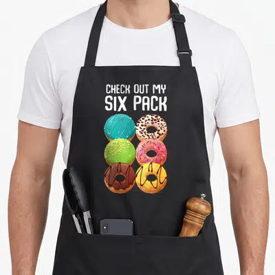 Gym donut apron - Six cartoon donuts in blue pink green yellow and brown below white text reading CHECK OUT MY SIX PACK — shown on a black cotton canvas apron against a white background