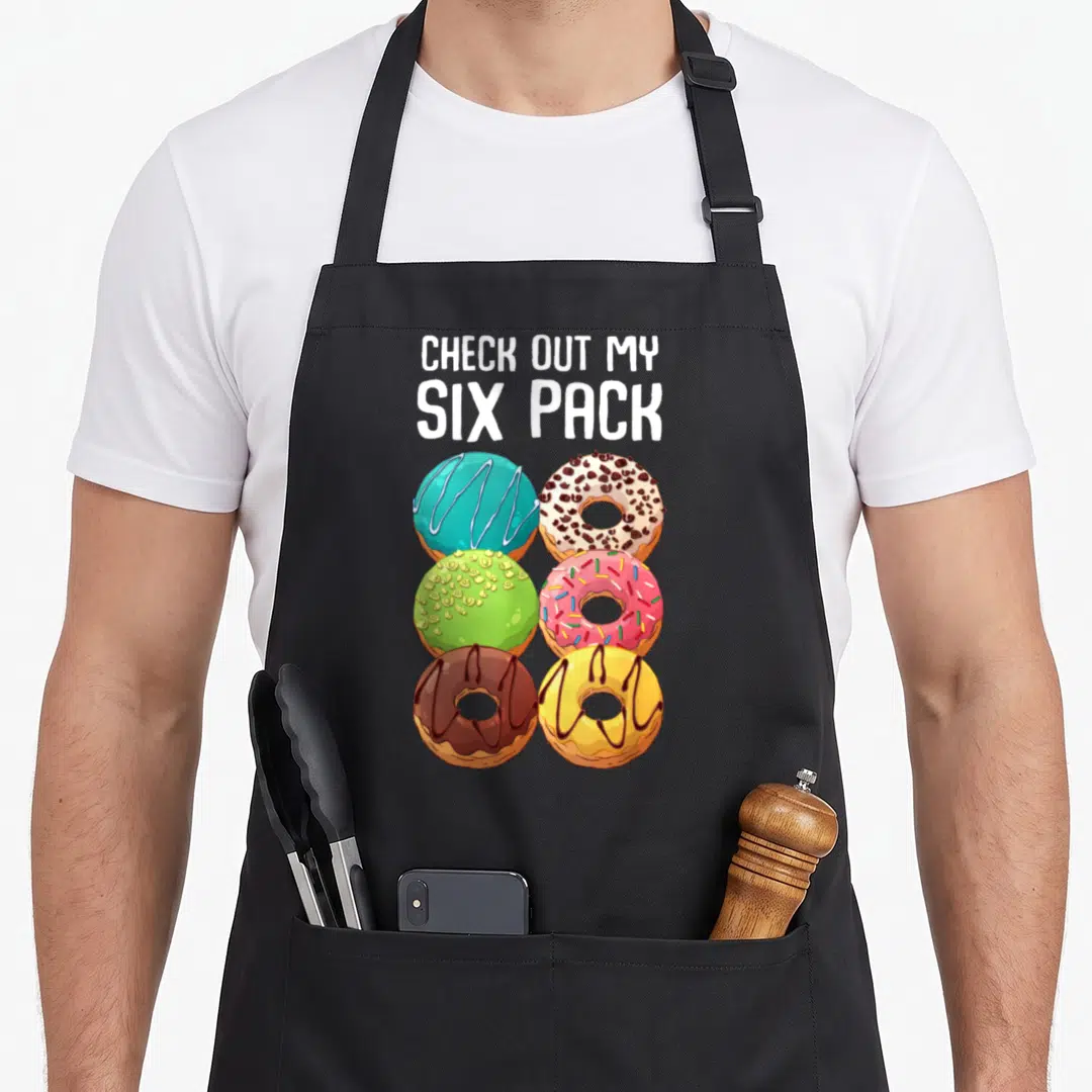 Gym donut apron - Six cartoon donuts in blue pink green yellow and brown below white text reading CHECK OUT MY SIX PACK — shown on a black cotton canvas apron against a white background