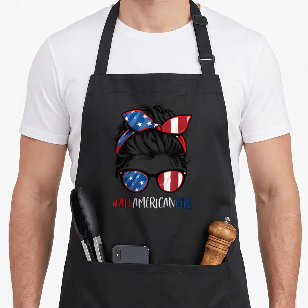 Funny flag patriotic 4th of july apron - Cartoon woman head with flag bow and sunglasses above #ALLAMERICANGIRL text — shown on a black cotton canvas apron against a white background