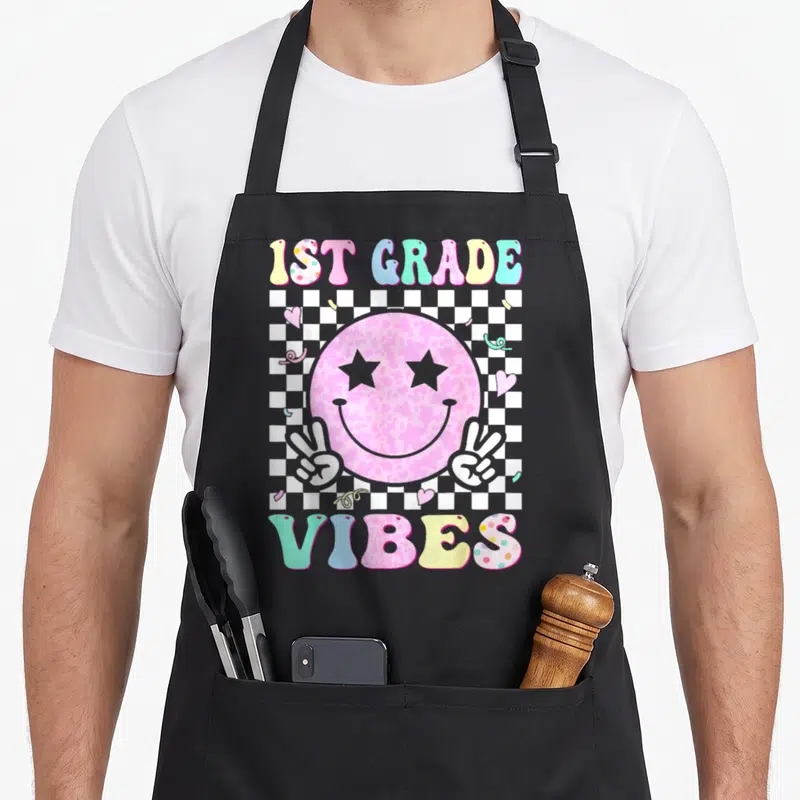 1st school grade vibes apron - Pink smiley face with star eyes and peace hands on checkerboard with hearts and confetti — shown on a black cotton canvas apron against a white background