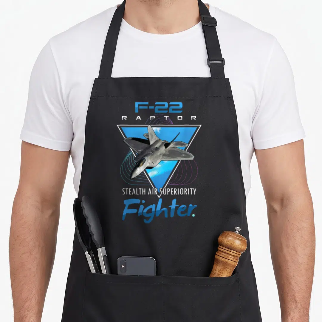 Raptor seasoned veteran age apron - Grey F-22 jet inside blue triangle with white clouds and stacked text reading F-22 — shown on a black cotton canvas apron against a white background