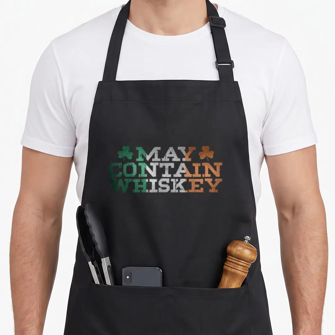 Contain funny irish aprons - Distressed May Contain Whiskey text in green white orange with shamrocks — shown on a black cotton canvas apron against a white background