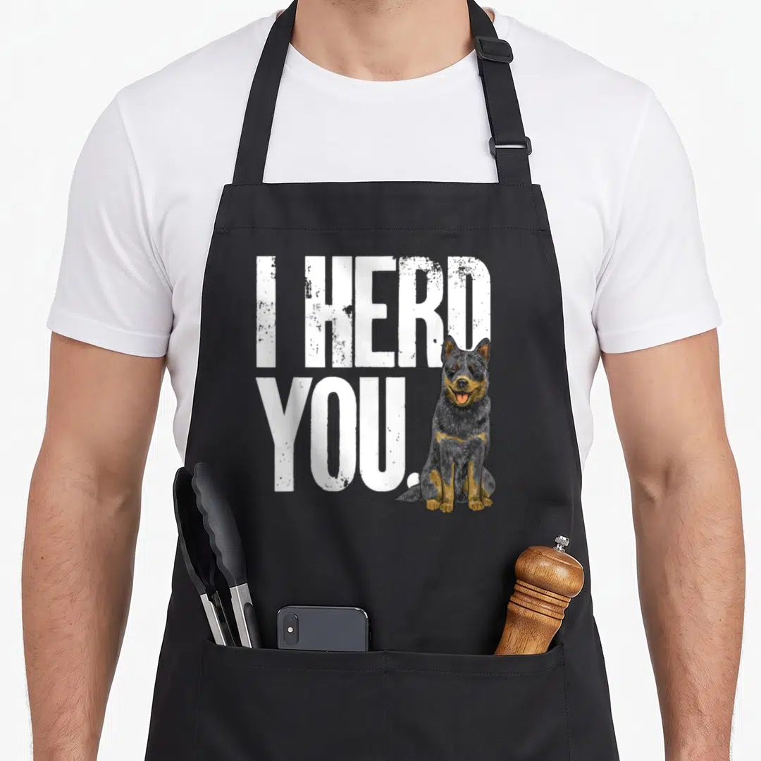 Heeler dog kitchen apron - I HERD YOU text with sitting blue heeler dog in white black and tan colors — shown on a black cotton canvas apron against a white background