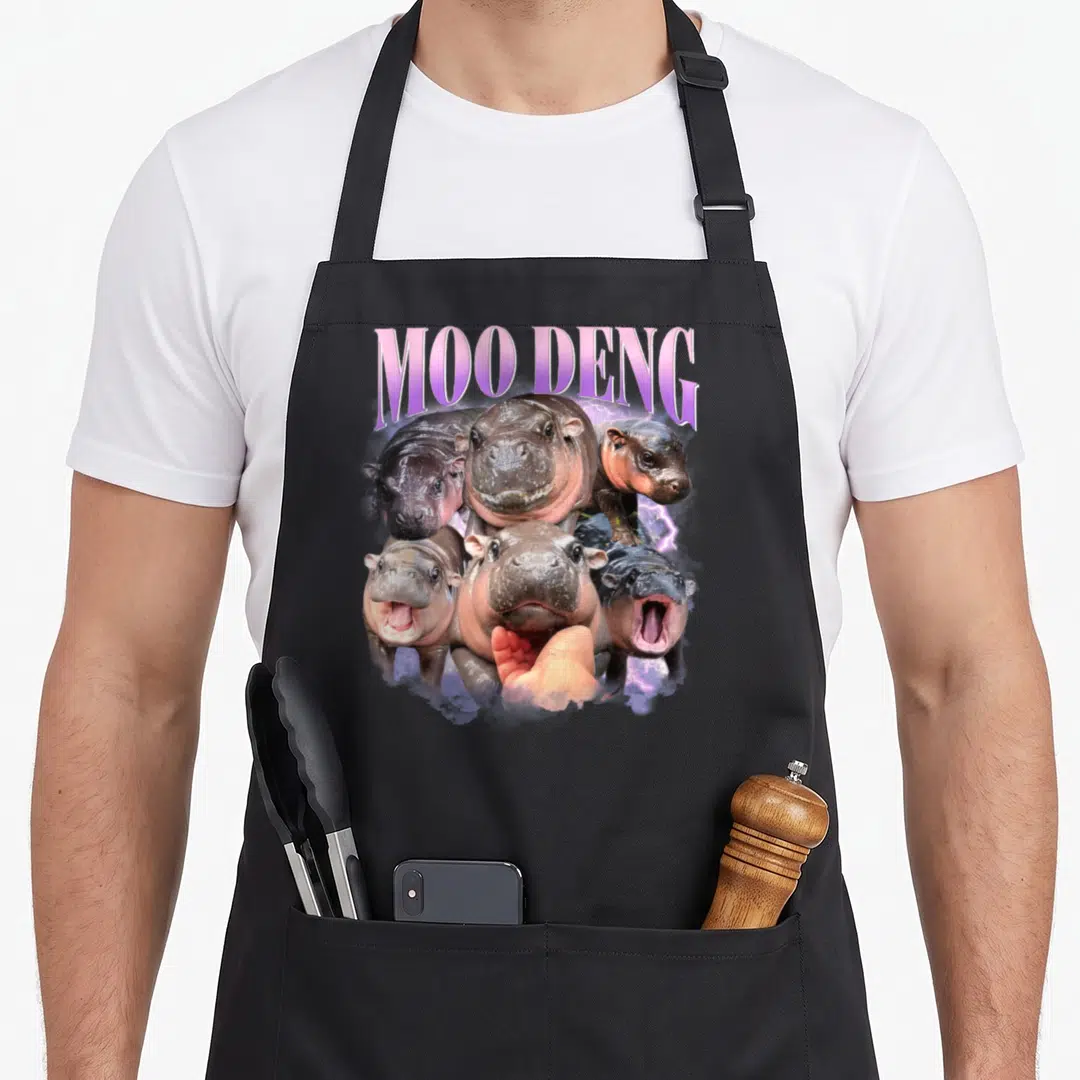 Moo family aprons - Six pygmy hippo heads under arched MOO DENG text with purple lightning — shown on a black cotton canvas apron against a white background