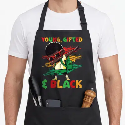 Dab african american aprons - Cartoon boy dabbing with YOUNG GIFTED & BLACK text and paint splashes — shown on a black cotton canvas apron against a white background