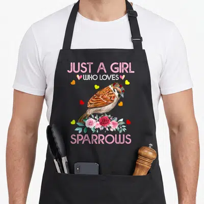 Cute sparrow bird apron - Watercolor sparrow with flower crown standing on pink roses surrounded by hearts and text — shown on a black cotton canvas apron against a white background