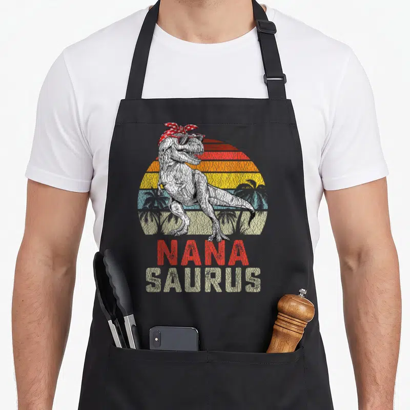 Rex nana aprons - T-Rex with sunglasses and bandana over striped sun with Nana Saurus text — shown on a black cotton canvas apron against a white background