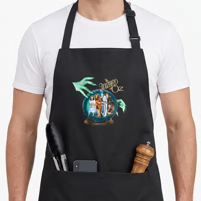 Crystal wizard of oz apron - The WIZARD of OZ text above crystal ball with four characters and two green hands — shown on a black cotton canvas apron against a white background