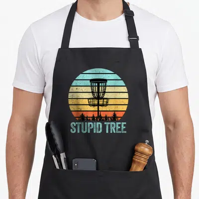 Stupid tree golf apron - Retro disc golf basket silhouette over striped sunset with stacked STUPID TREE text below — shown on a black cotton canvas apron against a white background