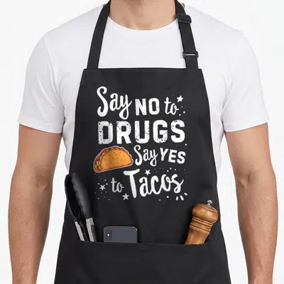 Ribbon taco apron - White text saying Say NO to DRUGS Say YES to Tacos next to an orange and red cartoon taco with stars — shown on a black cotton canvas apron against a white background