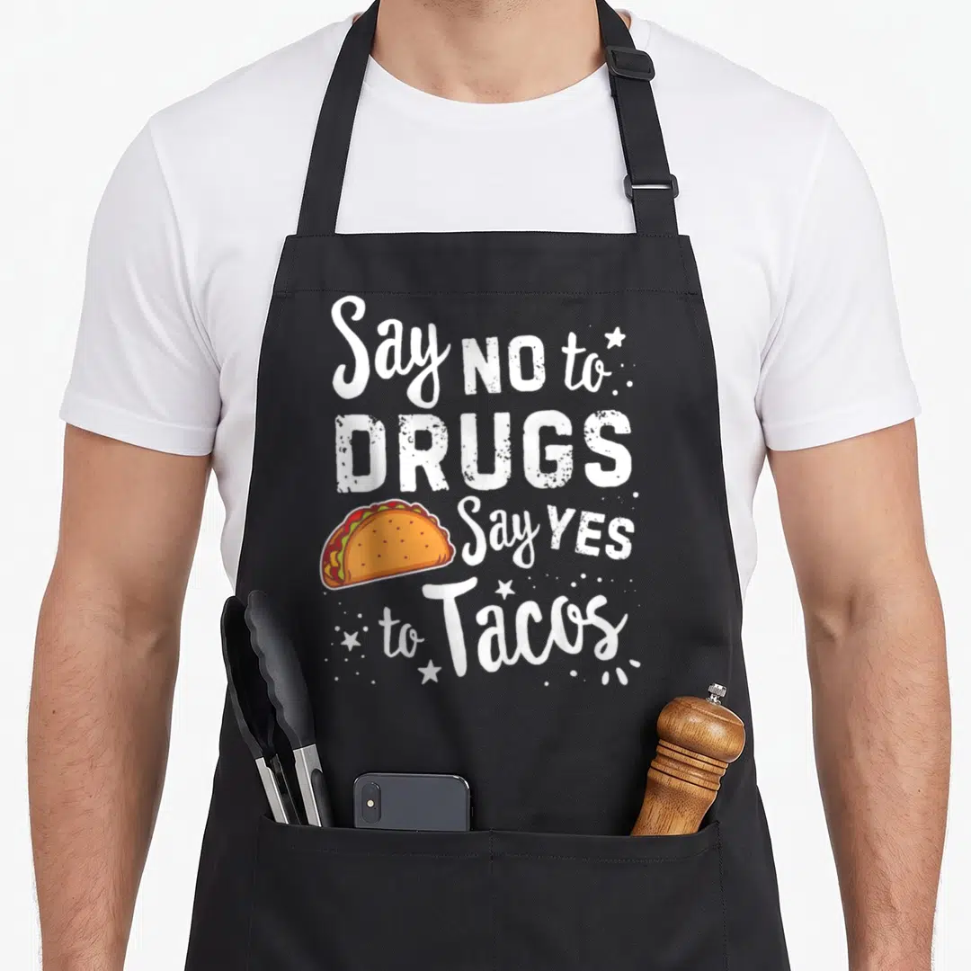 Ribbon taco apron - White text saying Say NO to DRUGS Say YES to Tacos next to an orange and red cartoon taco with stars — shown on a black cotton canvas apron against a white background