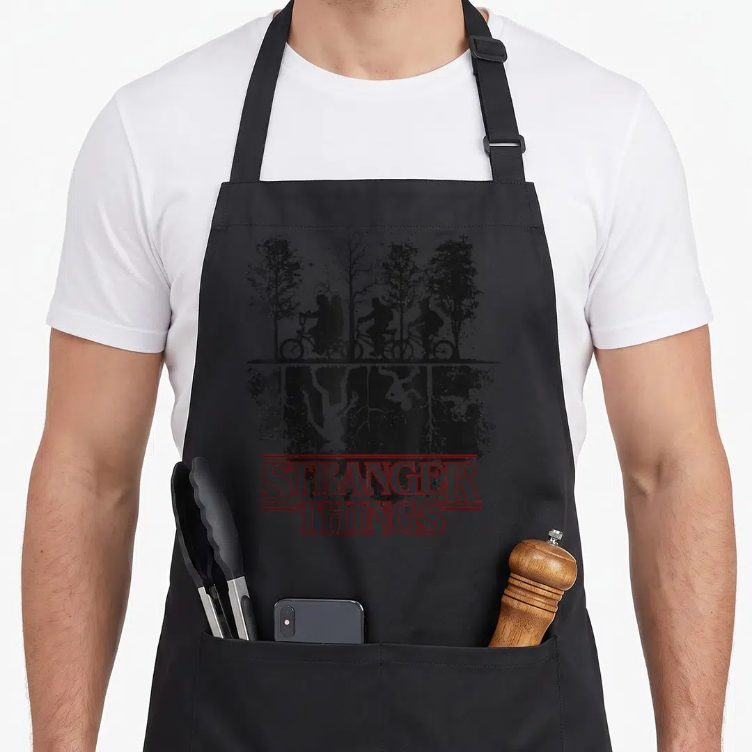Upside novelty apron - Black silhouette of kids on bikes above inverted monster roots with red STRANGER THINGS text — shown on a black cotton canvas apron against a white background