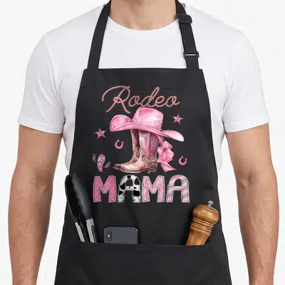 Rodeo mama cowgirl aprons - Watercolor brown boots pink hat cactus stars horseshoes and patterned MAMA text — shown on a black cotton canvas apron against a white background