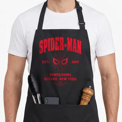Homecoming collegiate aprons - Red spider web with mask eyes and text reading Spider-Man Est 2017 Queens New York — shown on a black cotton canvas apron against a white background