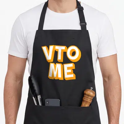 Associate swagazon apron - VTO ME text in white with orange outline stacked vertically — shown on a black cotton canvas apron against a white background