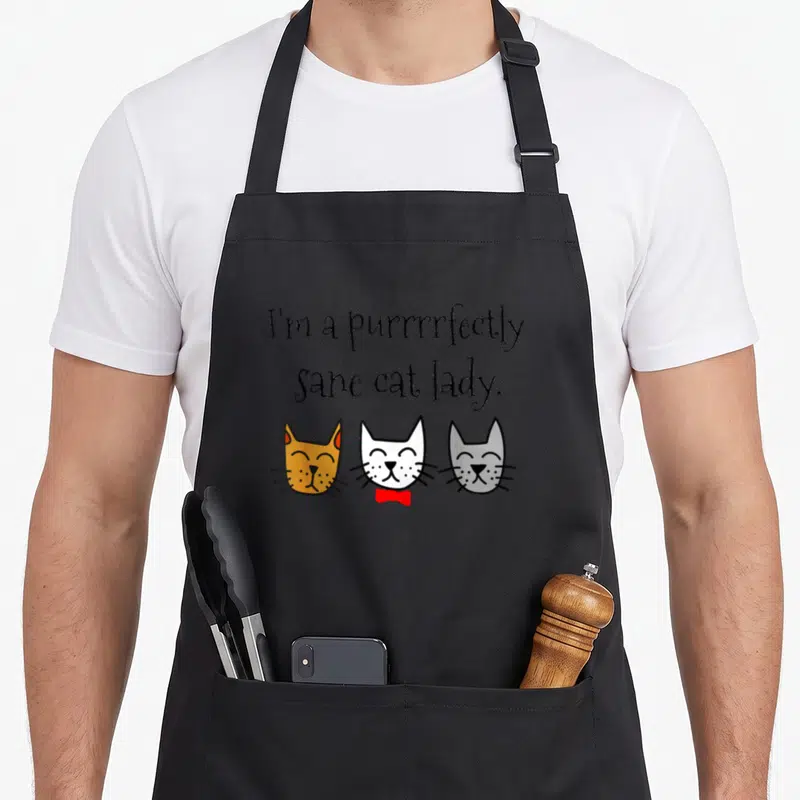 Perfectly crazy cat lady apron - Three cartoon cat heads below text reading I'm a purrrrfectly sane cat lady — shown on a black cotton canvas apron against a white background